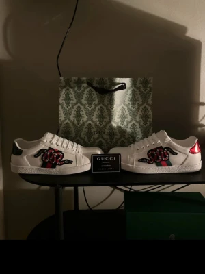 Gucci Ace sneakers  - Säljer ett par vita Gucci Ace sneakers i skinn med klassiska gröna och röda ränder på sidan och en broderad orm. Skorna har snörning, platt sula och rund tå. Baksidan har metallicröd och grön detalj. Snygg och ikonisk design som sticker ut.
