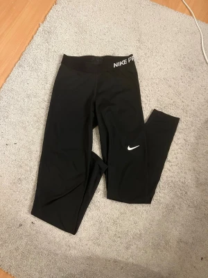 Svarta Nike Pro tights - Svarta Nike Pro tights med vit Nike-logga på benet och bred resår med Nike Pro-tryck i vitt. Snygg och sportig design som passar perfekt till träning. Materialet är stretchigt och följsamt för maximal komfort. Aldrig använda av mig men köpta på Vinted, finns spår av användning 