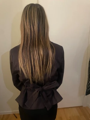 Svart kavaj med bälte och knappar - Snygg svart kavaj med två stora knappar framtill och ett brett bälte som knyts i ryggen. Jackan har klassisk krage och figurnära passform som ger en stilren siluett. Perfekt för dig som vill ha en chic och modern look.