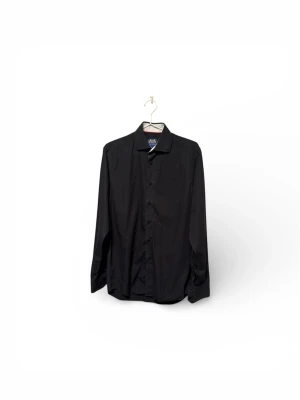 Svart skjorta från John Henric - Svart skjorta från John Henric med klassisk krage och knappar framtill. Skjortan har lång ärm och är tillverkad i mjuk bomull som känns skön mot huden. Perfekt för dig som gillar stilrena och enkla plagg med snygg passform.