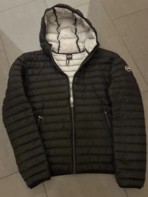 Colmar - Snygg svart pufferjacka från Colmar med svart foder och huva. Jackan har quiltad design, dragkedja framtill och två sidofickor med dragkedja. Perfekt för kalla dagar och har en modern, clean look med Colmar-logga på ärmen.