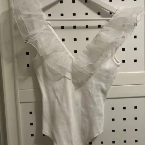 Vit ärmlös body med volang - Säljer en vit ärmlös body med djup v-ringning och stora, transparenta volangdetaljer över axlarna. Toppen har en elegant och unik look som sticker ut och passar perfekt till dig som vill ha något extra.❤️