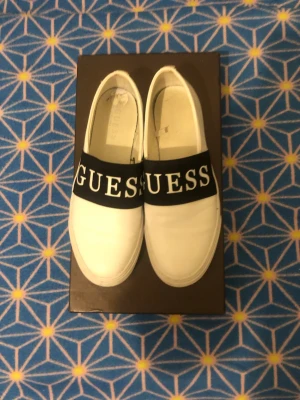 Vita slip-on sneakers från Guess - Säljer ett par vita slip-on sneakers från Guess med bred svart rem över foten där det står GUESS i stora vita bokstäver. Skorna har rund tå och platt sula med Guess-logga under. Snygg och enkel design som passar till jeans eller shorts.