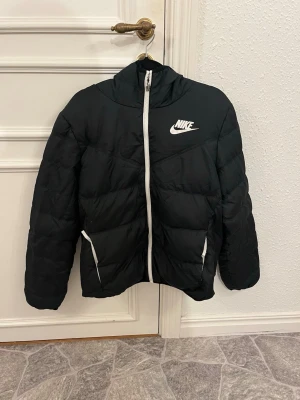 Svart pufferjacka från Nike, M - Säljer en svart pufferjacka från Nike i storlek M. Jackan har vit dragkedja, två fickor med vita detaljer och en hög krage. Klassisk Nike-logga på bröstet och skön passform. Perfekt för kyliga dagar och enkel att styla till det mesta.
