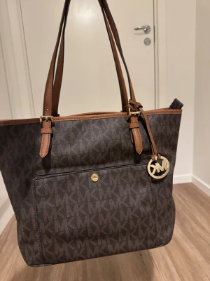 Brun axelväska från Michael Kors - Snygg brun axelväska från Michael Kors med MK-logga i mönstret och guldfärgad detalj framtill. Väskan har långa handtag i brunt skinn, flera fack och dragkedja upptill. Insidan är fodrad med ljus textil med Michael Kors-tryck. Perfekt storlek för allt du behöver.