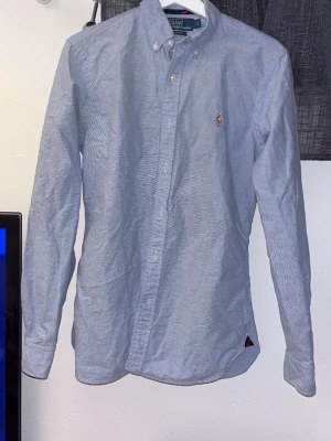 Blå slim fit skjorta från Polo Ralph Lauren - Säljer en stilren blå skjorta från Polo Ralph Lauren i slim fit-modell. Skjortan har klassisk button-down krage, lång ärm och knappar framtill. Materialet är mjuk bomull och den har den ikoniska broderade loggan på bröstet. Perfekt för dig som gillar clean och snygg stil.