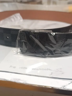 Svart rutig Louis Vuitton bälte - Säljer ett svart bälte från Louis Vuitton med klassiskt rutigt mönster och LV-logga på spännet. Bältet har en snygg matt finish och insidan är ljusbrun. Perfekt accessoar för att lyfta din outfit.