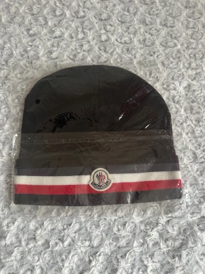 Svart mössa med röd-vit rand - Svart mössa från med klassisk rund form och bred uppvikt kant. Dekorerad med en röd och vit rand samt -logga framtill. Materialet är mjuk stickad bomull som känns skön mot huvudet. Perfekt för dig som vill ha en stilren och sportig look.