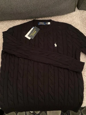 Svart kabelstickad tröja Polo Ralph Lauren - Svart kabelstickad tröja från Polo Ralph Lauren med klassisk rund hals och vit broderad logga på bröstet. Tröjan har långa ärmar och ribbade muddar vid hals, ärmslut och nederkant. Perfekt för dig som gillar stilrena och tidlösa plagg.