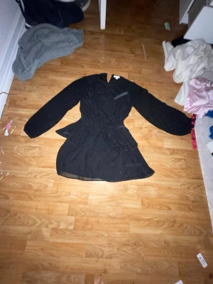 Svart glittrig kortklänning med volang - Säljer en svart kortklänning ifrån Gina tricot med glittriga prickar och volangdetaljer. Klänningen har långärmade transparenta ärmar och markerad midja som ger en snygg siluett. Perfekt för dig som gillar lite extra glam och vill sticka ut.endast använd 1 gång 