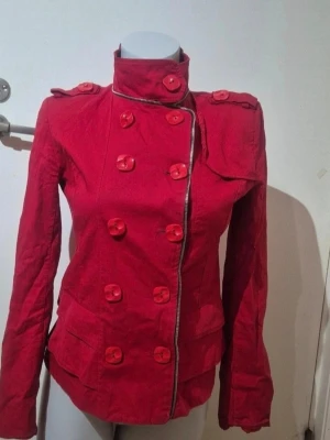Röd rock med knappar och hög krage - Snygg röd rock med dubbelknäppning, hög krage och dekorativa axelklaffar. Jackan har långa ärmar, markerad midja och coola detaljer som stora knappar och dragkedja i halsen.  jackan har en feminin, figurnära passform.Kort kappa, färgen är mindre rödaktig än på bilderna ❤️🌹🥰 Den har vissa tecken på slitage, den har använts flera gånger. Storlek L, se sista bilden.