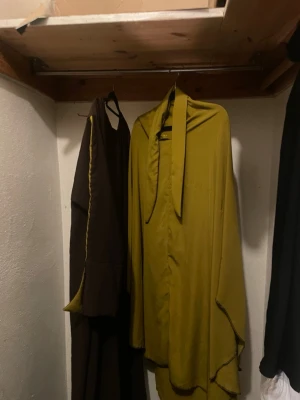 Långklänning i olivgrönt och brunt - Unik långklänning i olivgrönt och brunt med kontrasterande gulgrön insida och dekorativ söm längs ärmen. Klänningen har en lös passform, långa ärmar och en bred krage med knytband. Materialet känns mjukt och har ett lätt glansigt utseende.