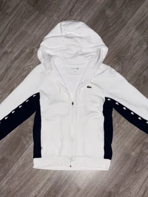 Lacoste Zip Hoodie - Vit Lacoste zip-hoodie med svarta detaljer längs sidorna och armarna samt små Lacoste-loggor längs ärmen. Broderad klassisk krokodil-logga på bröstet. Hel dragkedja framtill och huva med snörning.