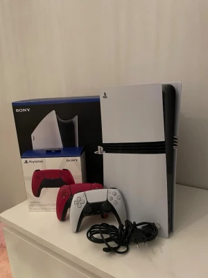 PlayStation 5 pro - Sony PlayStation 5 i mycket fint skick, komplett med originalkartong, två DualSense-kontroller (vit och röd) samt strömkabel. Konsolen och tillbehören ser ut att vara välvårdade utan synligt slitage eller defekter. Perfekt för gaming med topprestanda och modern design. KVITTO FINNS MED!