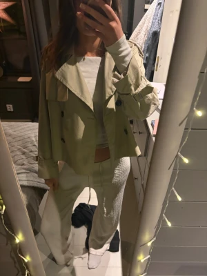 Kort beige trenchcoat med bälte - Säljer en kort och trendig beige trenchcoat med dubbelknäppning och snygga axeldetaljer. Jackan har bälte vid ärmsluten och bred krage, perfekt för lager-på-lager. Passformen är loose och jackan är i ett klassiskt trench-material.