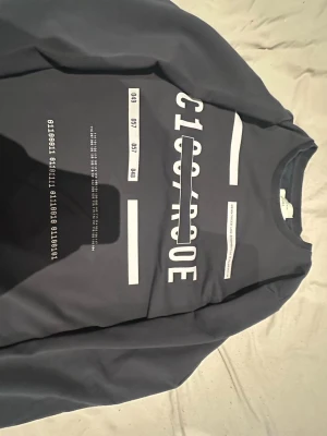 Mörkblå sweatshirt från Jack & Jones - Säljer en mörkblå långärmad sweatshirt från Jack & Jones Core med vit grafisk text och tryck på framsidan. Tröjan har rund hals, ribbade muddar och är gjord i mjuk bomull. Perfekt för chill dagar och enkel att matcha med jeans eller joggers.