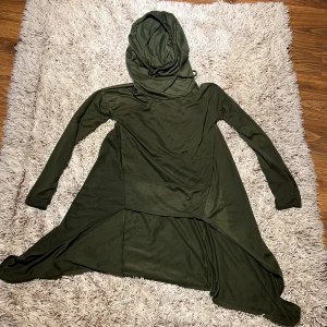 Olivgrön lång hoodie med fickor - Säljer en olivgrön lång hoodie i storlek M.  Hoodien har huva, långa ärmar och praktiska fickor framtill.  Materialet är mjukt och känns som bomull.  Den har en cool asymmetrisk skärning nedtill som ger en unik look. Den är i använt skick. Från Wish