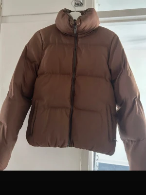 Brun pufferjacka med dragkedja - Säljer en brun pufferjacka med hög krage och dragkedja framtill. Jackan har en boxig form och är vadderad för extra värme. Perfekt för kalla dagar och har två fickor på sidorna. Enkel och stilren design som är lätt att matcha.