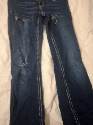 Mörkblå jeans med kontrastsömmar - Säljer ett par mörkblå jeans med vita kontrastsömmar och slitna detaljer på benen. Modellen har klassiska fem fickor och dekorativa knappar. Jeansen har en rak passform och är tillverkade i jeansmaterial. Perfekt för dig som gillar en edgy stil.