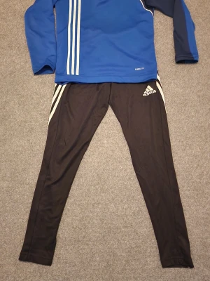 Adidas tracksuit - Blå och vit adidas tröja med adidas svarta byxor för barn i fint skick. Byxorna har dragkedja på båda fickor samt dragkedja längst ner vid benen. Storlek på tröjan 152 eller 11-12 år. Och byxorna 11-13 år. 
