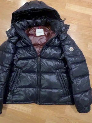 Svart Moncler dunjacka med huva - Snygg svart dunjacka från Moncler med glansig finish och huva. Jackan har dragkedja framtill, två sidofickor med dragkedja och klassisk Moncler-logga på ärmen. Insidan är vinröd och materialet är polyester. Perfekt för kalla dagar.