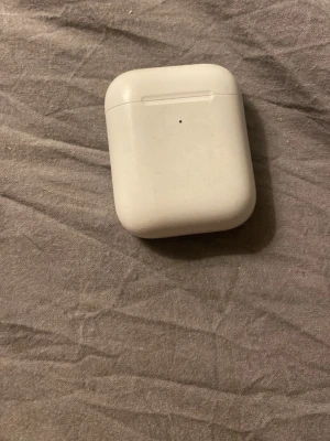 Apple AirPods med laddningsetui - Säljer ett par Apple AirPods med vitt laddningsetui. Etuiet är kompakt, har rundade hörn och är gjort i blank plast. Perfekt för dig som vill ha trådlösa hörlurar med smidig laddning och enkel anslutning till din iPhone eller andra Apple-enheter.