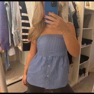 Blåvit randig axelbandslös topp - Säljer en blåvit randig axelbandslös topp med knappar framtill. Toppen har bandeauformad överdel och lätt utställd passform nedtill. Perfekt för dig som gillar en fräsch och somrig look.