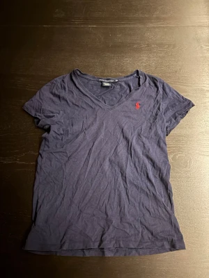 Marinblå v-ringad t-shirt Ralph Lauren - Snygg marinblå t-shirt från Ralph Lauren Sport med v-ringning och korta ärmar. Tillverkad i 100% bomull och har den klassiska röda loggan broderad på bröstet. Perfekt basplagg för en avslappnad stil.