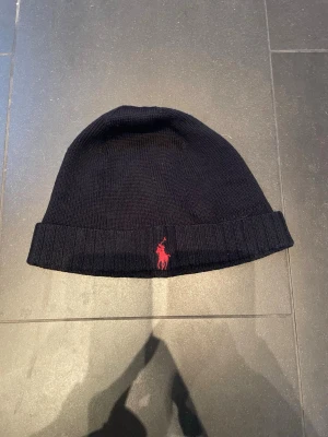 Svart mössa från Polo Ralph Lauren - Klassisk svart ribbstickad mössa från Polo Ralph Lauren med uppvikt kant och röd broderad logga framtill. Perfekt accessoar för kyliga dagar och enkel att matcha med olika stilar. Tillverkad i mjukt material för skön komfort.