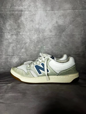 Vita New Balance sneakers med blå detalj - Snygga vita sneakers från New Balance med blå logga på sidan och ljusgrå mockadetaljer. Skorna har klassisk snörning, rund tå och platt sula. Perfekta för dig som gillar stilrena och sportiga sneakers med retrovibbar.