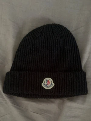 Svart ribbstickad mössa från Moncler - Säljer en svart ribbstickad mössa från Moncler med uppvikt kant och klassiska Moncler-loggan framtill. Perfekt för dig som vill ha en stilren och tidlös accessoar till höst och vinter.