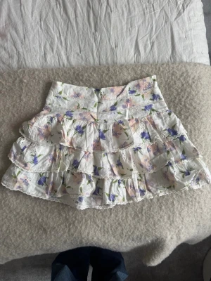 Vit volangkjol från Zara med blommor – storlek M - Säljer en vit volangkjol från Zara i storlek M med ett fint blommönster. En feminin och somrig kjol med vacker volangdetalj som ger ett luftigt och elegant fall. Perfekt för både vardag, semester och festligare tillfällen. Lätt att styla med linne, blus eller stickad tröja. 