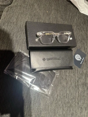Transparent Chrome Hearts glasögon - Säljer ett par riktigt snygga transparenta glasögon från Chrome Hearts. Bågarna är i klar plast med coola silvriga detaljer vid gångjärnen och logga på skalmarna. Medföljer original svart fodral och putsduk. Perfekt accessoar för dig som vill sticka ut.