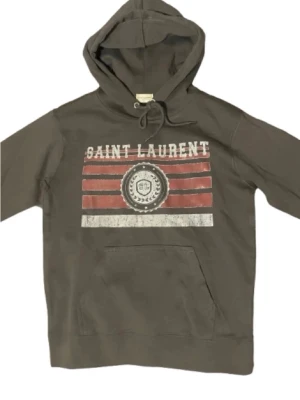 Grå Saint Laurent hoodie med tryck - Snygg grå hoodie från Saint Laurent med huva och dragsko. Framsidan har ett vintageinspirerat tryck med röda och vita ränder samt texten 'SAINT LAURENT'. Stor känguruficka framtill. Tillverkad i mjuk bomull för skön känsla.