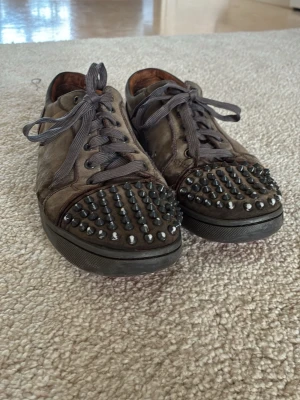 Christian Louboutin With Spikes - Unika bruna sneakers i skinn med coola svarta nitar över tån och klassisk röd sula. Skorna har snörning och en rund tå, vilket ger en edgy vibe. Perfekta för dig som vill sticka ut med din stil.