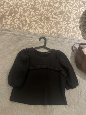 Svart blus med puffärm från Zara - Trendig svart blus från Zara med puffiga trekvartsärmar och volangdetalj framtill. Baksidan har tre dekorativa knappar i silver ( Har en defekt vilket är en sminkfläck som är minimal på är knappen högst upp på baksidan.