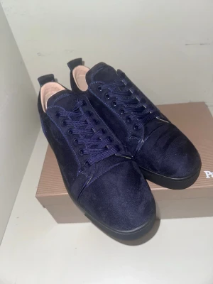 Mörkblå mocka sneakers  - Säljer ett par mörkblå sneakers i mjuk mocka med matchande snörning och svarta platta sulor. Skorna har den klassiska röda undersidan och en stilren, rund tå. Perfekta för dig som gillar exklusiva detaljer och snygg design. Kvitto, dustbag, låda, kort och papper ingår!