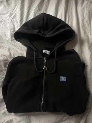 Svart hoodie från Les Deux - Snygg svart hoodie från Les Deux med dragkedja framtill och justerbar huva. På bröstet sitter en liten blå patch med vita bokstäver 'II'. Perfekt för en chill och stilren look. Tillverkad i mjukt bomullsmaterial för extra komfort.