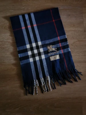 Mörkblå rutig halsduk - Snygg mörkblå halsduk med klassiskt rutmönster i vitt, svart och rött. Halsduken har fransar i ändarna och en broderad logga med ryttare. Perfekt accessoar för att lyfta din outfit.