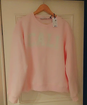 Ljusrosa sweatshirt med CALI-tryck - Säljer en ljusrosa sweatshirt med texten 'CALI' i vitt framtill. Tröjan har rund halsringning, långa ärmar och ribbade muddar vid ärmslut och nederkant. Perfekt för en avslappnad och trendig look. Materialet känns mjukt och bekvämt.