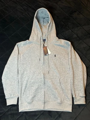 Grå zip-hoodie Polо Ralph Lauren - Snygg grå hoodie från Polo Ralph Lauren med dragkedja framtill, huva med snörning och klassisk logga broderad på bröstet. Tillverkad i mjuk bomullsmix och har två fickor fram. Perfekt för en chill och stilren look.