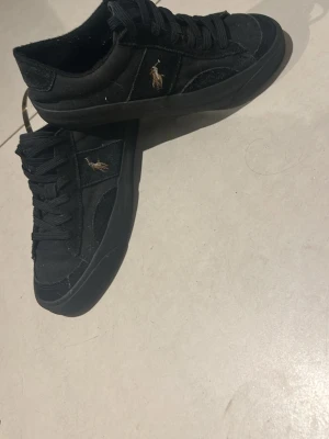 Svarta sneakers från Polo Ralph Lauren - Stilrena svarta sneakers från Polo Ralph Lauren med klassisk låg profil och guldfärgad logga på sidan. Skorna har snörning och är tillverkade i mocka och textil med platt sula och rund tå. Perfekta för en avslappnad och trendig look. Det är bara att höra av er vid frågor eller mer bilder på skorna 