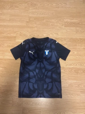 Puma Malmö FF fotbollströja - Snygg och funktionell fotbollströja från Puma, i en mörkblå/svart design med stilrent mönster. Tröjan har Malmö FF:s klubbmärke på bröstet och är tillverkad i Pumas dryCELL-material som andas och transporterar bort fukt – perfekt för träning eller match.