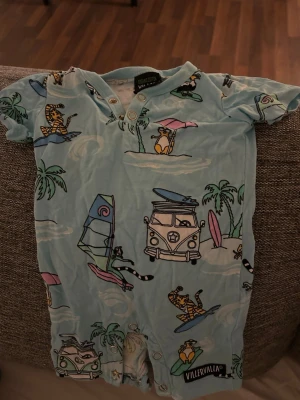 Blå body med surfmönster VillerValla - Supersöt ljusblå body från VillerValla med tryck av surfande tigrar, palmer, bussar och vindbrädor. Bodyn har tryckknappar fram och i grenen för enkel påklädning. Tillverkad i mjuk bomull, perfekt för sommarens äventyr.