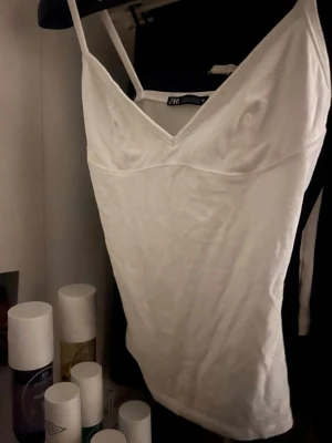 Vit linnetopp från ZARA - Säljer en enkel och stilren vit linnetopp från ZARA med smala axelband och v-ringad hals. Toppen är figurnära och har en mjuk, lätt stretchig känsla. Perfekt att matcha med jeans eller kjol för en clean look. Ej använd