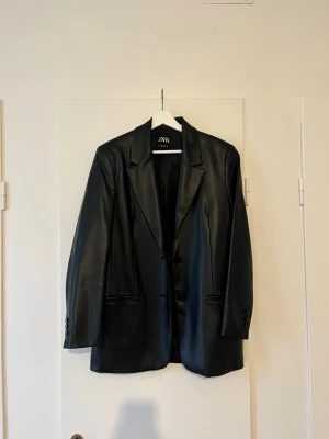 Svart kavaj i skinnimitation från Zara - Stilren svart kavaj från Zara i skinnimitation med klassisk krage och knäppning framtill. Kavajen har raka axlar, långa ärmar och fickor fram. Fodrad med 100% återvunnen polyester för extra komfort. Perfekt för en trendig och edgy look.
