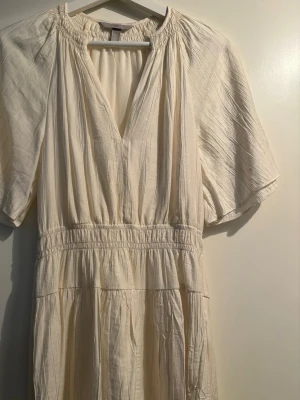 Ljus kortklänning med v-ringning - Luftig och ljus kortklänning i mjuk bomull med v-ringad hals och korta ärmar. Klänningen har resår i midjan för en bekväm passform och ett lätt rynkat fall. Perfekt för varma dagar och enkel att styla.