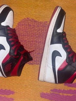 Air Jordan 1 svart/röd/vit sneakers - Säljer ett par Air Jordan 1 sneakers i svart, rött och vitt. Klassisk high-top modell med röd snörning, svart swoosh och ikonisk Air Jordan-logga på sidan. Ovandelen är i skinn och sulan är vit med röd kant. Perfekta för dig som gillar streetwear.