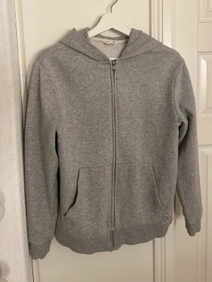 Grå zip hoodie med huva - En basic grå hoodie med dragkedja framtill och två fickor. Hoodien har en klassisk huva och ribbade muddar vid ärmar och nederkant. Perfekt för chill dagar eller när du vill ha en enkel och stilren look. Materialet känns mjukt och bekvämt.