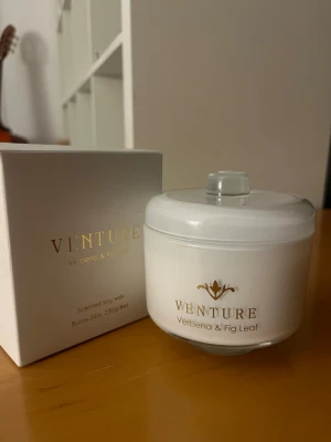 Doftljus Venture Verbena & Fig Leaf - Snyggt vitt doftljus från Venture med doft av verbena och fikonblad. Ljuset är i en rund glasburk med lock och har två vekar. Levereras i stilren vit kartong med gulddetaljer. Tillverkat av sojavax och väger 250g.
